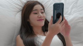 top view. Young asian girl lying on bed and playing smart phone. Beautiful woman using cellphone to chat with friends and play social media. Lifestyle relaxation and technology concept
