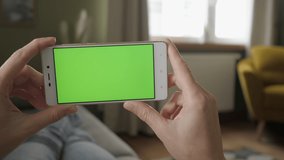 Young Woman Home Lying on a Couch with Green Screen Smartphone in Horizontal Mode. Girl Using Touchscreen Mobile Phone. Girl Using Smartphone, Browsing Internet, Watching Video Content, Blogs. POV.