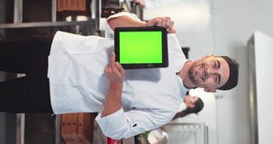 Vertical concept. Portrait of young cheerful smiling professional male chef standing in restaurant kitchen in uniform showing tablet with chroma key. Device with green screen, tech culinary