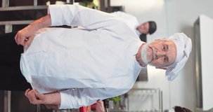 Vertical video of Caucasian grey-haired professional male chef putting medical mask on face standing in own restaurant kitchen and looking at camera before working process. Portrait, workflow behind