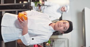 Vertical video of young pretty Asian cheerful professional woman chef looking at camera and smiling standing in restaurant kitchen throwing in air yellow bell pepper. Portrait of happy female cook