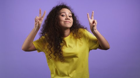 Pretty woman with curly hair showing with hands and two fingers air quotes gesture, bend fingers isolated on violet background. Not funny, irony and sarcasm concept.