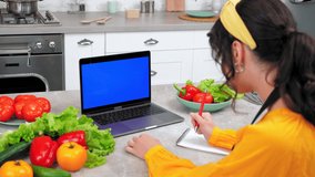 Blue screen mock up chroma key monitor laptop: Woman housewife in kitchen watch online cooking course listen teacher tells chef writes culinary recipe in notebook, study remote video call computer