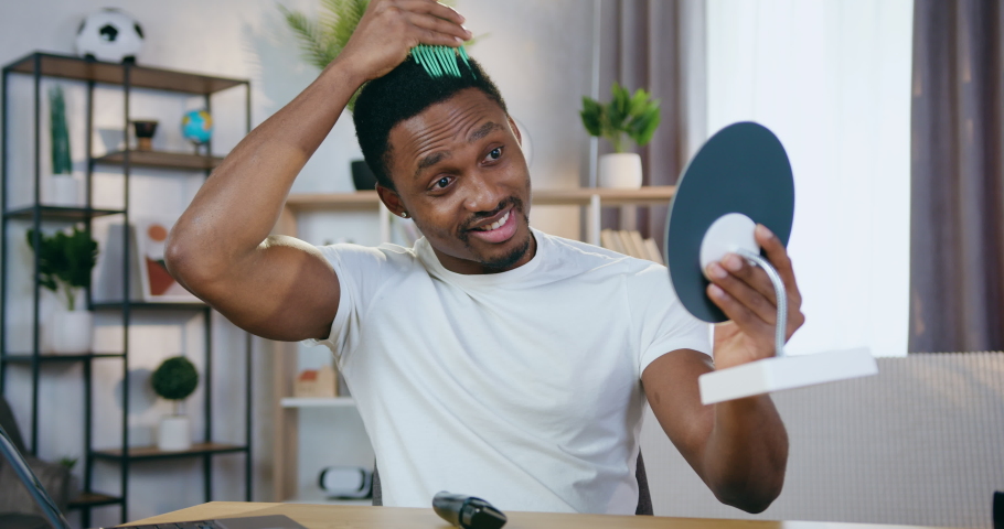 Likable joyful confident young black-skinned guy looking in small mirror and combing his hair and beard with special comb,slow motion