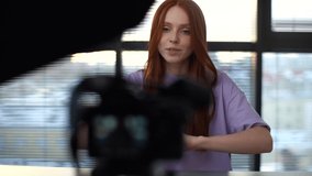 Close-up of smiling young woman blogger turns on camera and recording vlog on camera sitting on background of window. Happy redhead female vlogger record online video course at home office.