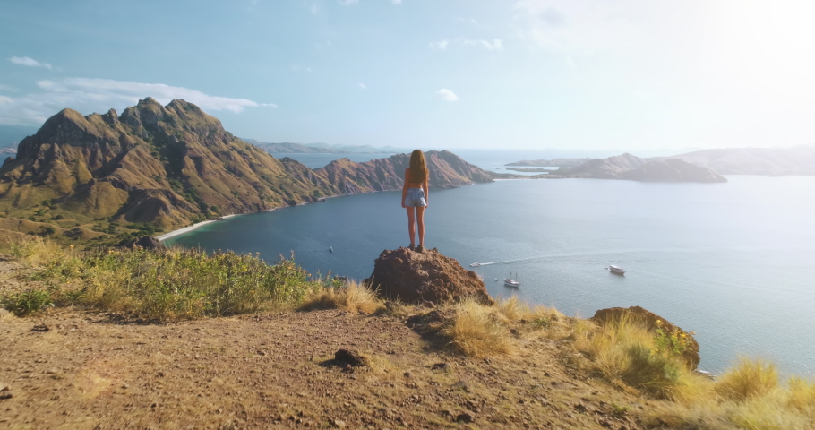 Girl rising hands on mountain at ocean harbor aerial view. Summer nature seascape with water transport. Woman fills freedom. Ship, yachts at sea shore. Travel lifestyle. Komodo Island, Indonesia, Asia Royalty-Free Stock Footage #1071316132