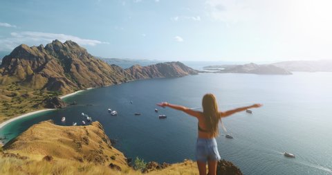 Girl rising hands on mountain at ocean harbor aerial view. Summer nature seascape with water transport. Woman fills freedom. Ship, yachts at sea shore. Travel lifestyle. Komodo Island, Indonesia, Asia