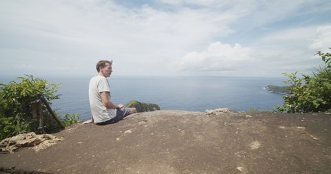 Low dolly shot of a male tourist sitting above the T-Rex Bay peninsula with reveal of the Kelingking beach in Bali, Indonesia Man sitting on a cliff lookout point above the tropical beach