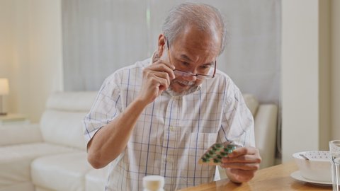 Asian Senior elderly male patient on eating table at nursing home care. Older man wearing eyeglasses and look to medicine pill in blister pack, read prescription and content. Medical insurance concept