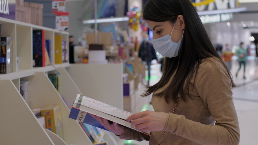 Long haired lady in disposable mask reads encyclopedia choosing present standing in bookstore
