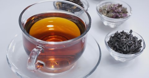 Drop slowly falls into a glass cup of tea. Around there are small bowls with different types of leaf tea. White background. 