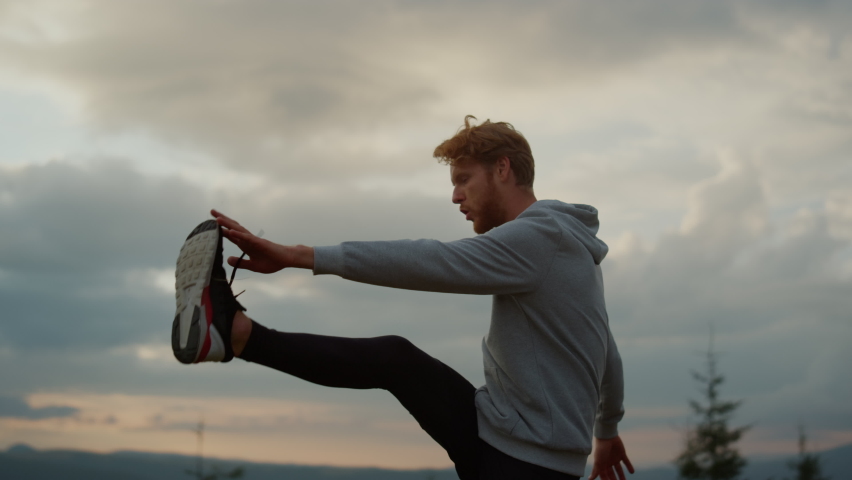 Male jogger warming legs before cardio workout in mountains. Active athlete lifting legs up to hands. Redhead man enjoying summer outdoor activity at sunset