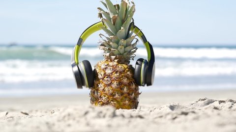 Funny pineapple in headphones, sandy ocean beach, blue sea water waves, California pacific coast, USA. Tropical summer exotic fruit enjoying vacations and music in paradise. Ananas sunbathing on shore