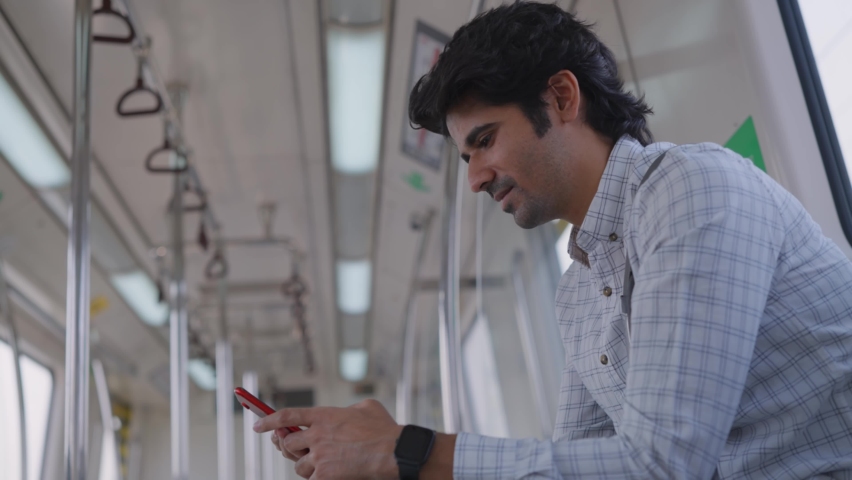 A young smailing attractive Indian male corporate employee in formal wear and sling bag is typing a text message on a mobile phone on a moving modern city metro train while going to the office  Royalty-Free Stock Footage #1071344335