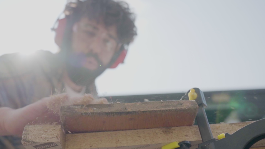 Carpenter blowing sawdust away from handcrafted wood plank on workbench outside the garage atelier. Artisan working wearing safety protections . Build home creative ideas, slow motion blurred subject | Shutterstock HD Video #1071344605