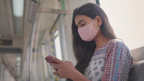 Close shot of a young attractive Indian female sitting on a moving city metro train with a protective mask on her face and typing a text message on a mobile phone during COVID 19 epidemic or pandemic 