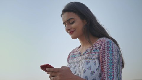 Close shot of a young beautiful Indian female using a mobile phone to type a text message with a smile on her face standing on top of a building terrace with a cityscape in the background