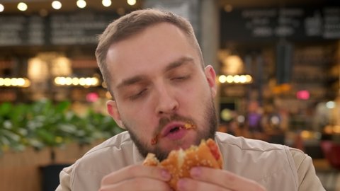 Unshaven caucasian man eats meat burger with pleasure, bites burger with his mouth, man casually eats junk food from fast food, smears his face with ketchup sauce on background of fast food restaurant