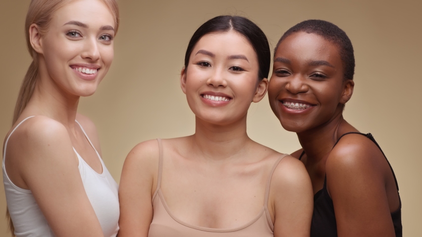 Studio portrait of three happy multiethnic ladies laughing to camera, enjoying friendship, hugging over beige background, slow motion Royalty-Free Stock Footage #1071348910