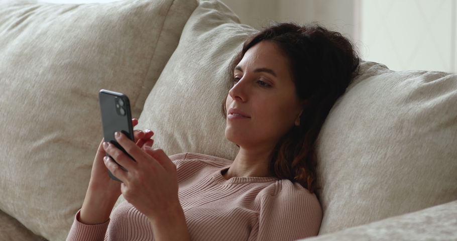Single beautiful relaxed woman lying leaned on sofa cushion use smartphone spend day off at home makes delivery order, enjoy easy comfort usage of modern tech. E-date, ecommerce, internet user concept Royalty-Free Stock Footage #1071353689