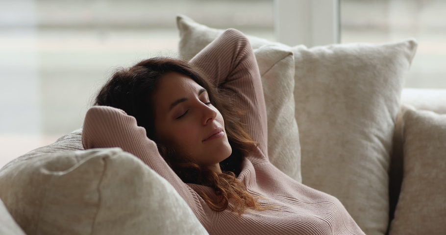 Attractive young female relaxing indoors leaned on sofa cushions. Woman breath fresh conditioned air folded hands behind head lying on cozy soft couch in living room. Meditation, rest, leisure concept Royalty-Free Stock Footage #1071353734