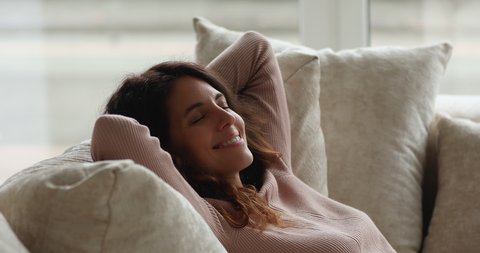 Attractive young female relaxing indoors leaned on sofa cushions. Woman breath fresh conditioned air folded hands behind head lying on cozy soft couch in living room. Meditation, rest, leisure concept