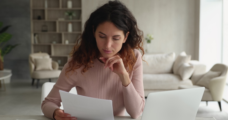 Attentive busy businesswoman sit at desk work holding documents analyzing information comparing data look at laptop screen. Growing successful small business owner, finances and sales research concept