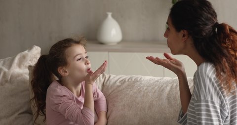 Little stuttering girl involved in speaking pronunciation lesson with female speech therapist seated on sofa at home. Training exercise for children to help with speech and language problems concept