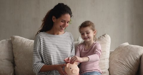 Thrifty kid girl puts coins into piggy bank while sit on couch with mom. Latina mother teach little daughter count and save money. Family having savings for future needs, insurance, investment concept