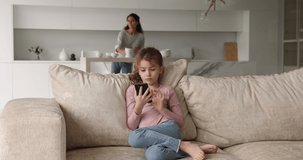 On foreground little girl sit on sofa having fun using smartphone while her young mother housewife do housework household chores get living room details in order. Family spend weekend at home concept