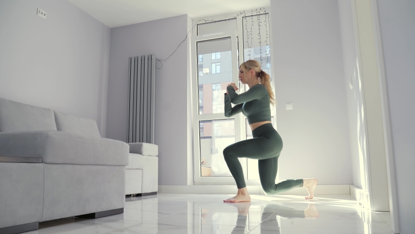 Young fitness woman doing squat exercises with lunges from leg to leg. Fit Female is a workout at home in the living room. Athletic attractive blonde trains muscles in the apartment indoors | Shutterstock HD Video #1071362284
