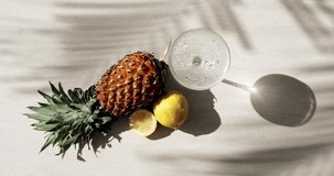 Close-up top-view of pineapple, lemon, palm tree shadows and a glass of soda in which a slice of lemon falls and creates a splash. High quality 4k footage
