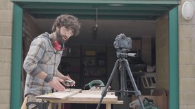 Young male craftsman recording video on reflex camera to show how to work and take measurement on wood plank outside garage shop. Personal channel or blog online and social marketing icons. Real-time - Powered by Shutterstock - Get 15% off with code: PIKWIZARD15