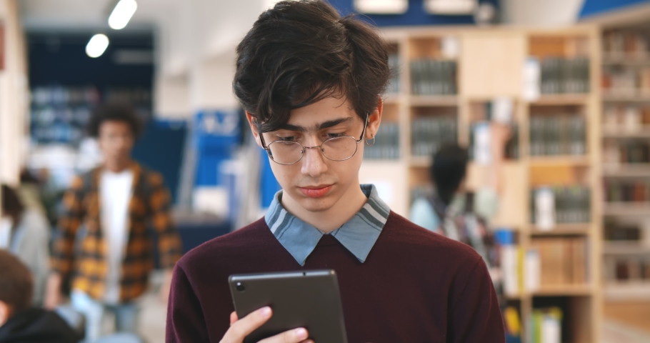 Male college student using digital tablet standing in library. Portrait of young man in eyeglasses reading ebook in tablet pc studying in university library Royalty-Free Stock Footage #1071373843