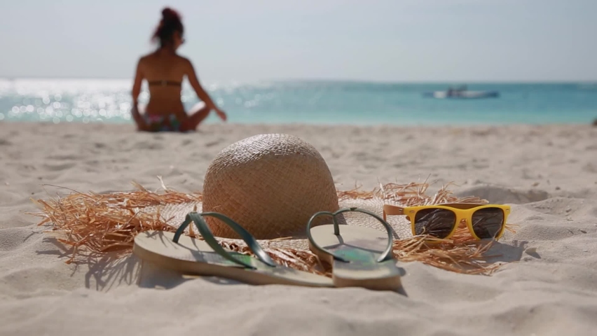A hat with large brim, flip-flops and yellow sunglasses lie on the beach on the sand, against the background of the sea and the silhouette of a girl sitting on the sand, the girl is out of focus Royalty-Free Stock Footage #1071375769