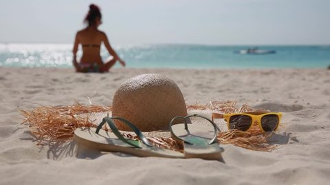 A hat with large brim, flip-flops and yellow sunglasses lie on the beach on the sand, against the background of the sea and the silhouette of a girl sitting on the sand, the girl is out of focus