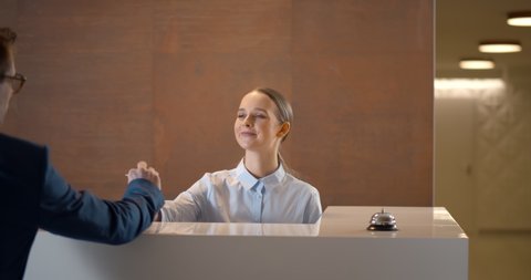 Young businessman with suitcase signing out of hotel leaving card key at reception. Smiling woman receptionist taking room key from guest leaving hotel