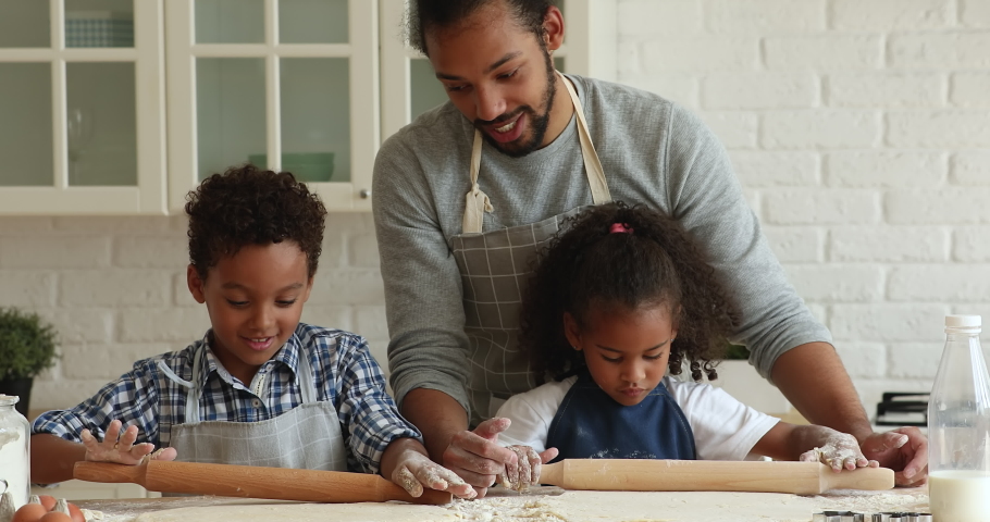 Smiling young caring afro father teaching helping little happy preschool son and daughter rolling out dough for handmade pastry, developing kids skills, enjoying cooking together on weekend at home. Royalty-Free Stock Footage #1071379567