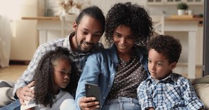 Smiling young african american mother holding smartphone in hands, showing funny video to happy husband and joyful little cute kids, posing for photo together or using apps indoors, tech addiction.