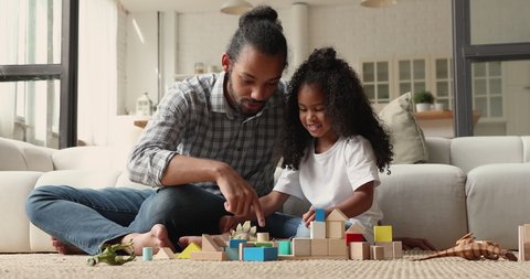 Caring young affectionate loving african american father playing toys with little cute biracial child daughter, sitting together on floor in modern living room, talking chatting communicating at home.