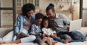 Addicted to technology young african american multiracial family couple sitting with little son and daughter on couch, involved in using different gadgets, playing mobile games, entertaining at home.