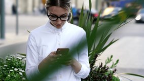 Caucasian female blogger in optical spectacles checking mobile notification for installing app, millennial woman browsing wireless website on cellular technology using 4g internet in city
