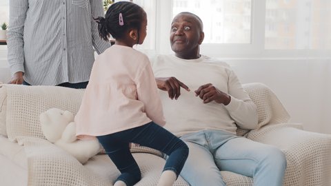 African little schoolgirl child girl talking with elderly man grandfather mature father sitting on couch afro black dad open eyes wide surprised from conversation unrecognizable woman stands near sofa