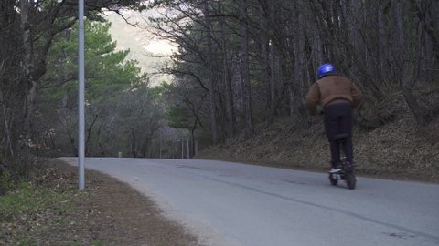 A driver wearing a protective helmet and a powerful electric scooter drives along a forest road. A man on an electric scooter in the forest on a concrete road quickly drives away from the camera