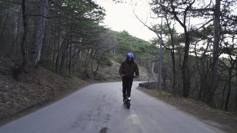 A driver wearing a protective helmet and a powerful electric scooter drives along a forest road. A man on an electric scooter in the forest rides wagging along the road
