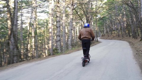 A driver wearing a protective helmet and a powerful electric scooter drives along a forest road. A man on an electric scooter rides a long distance in the forest along the road with his back to the