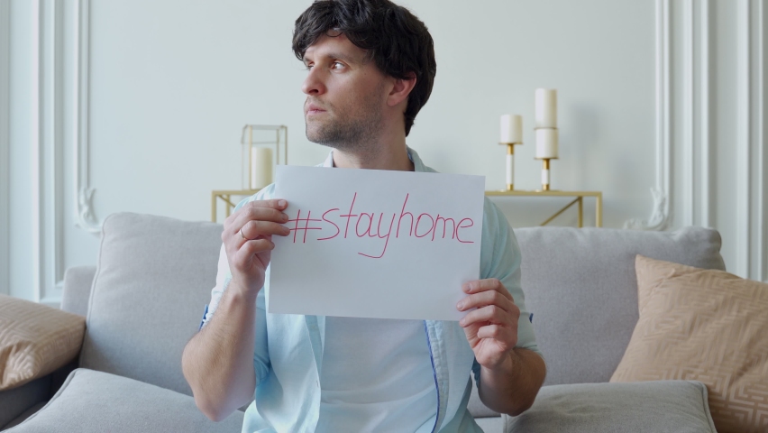 Man sitting on a sofa warns of coronavirus disease holds a placard stay at home Royalty-Free Stock Footage #1071394318