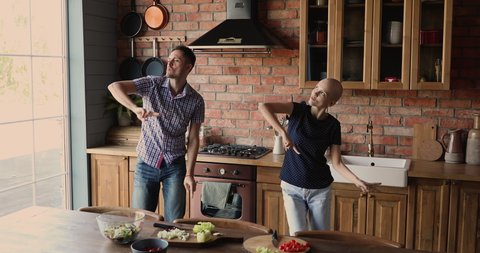 Happy young man dancing with energetic sincere hairless after chemotherapy wife, having fun celebrating oncology remission, distracted from cooking enjoying spending leisure time together at home.