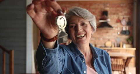 Overjoyed sincere middle aged senior woman holding keys in hands, showing to camera. Happy excited elderly female retired homeowner celebrating purchasing own dwelling, real estate service concept.