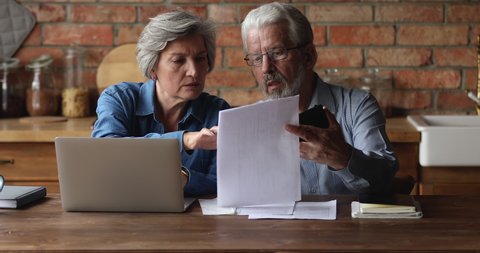 Serious older husband showing banking paper document bills taxes to confused elderly wife, sharing financial problems at home. Stressed middle aged family couple calculating expenses together indoors.